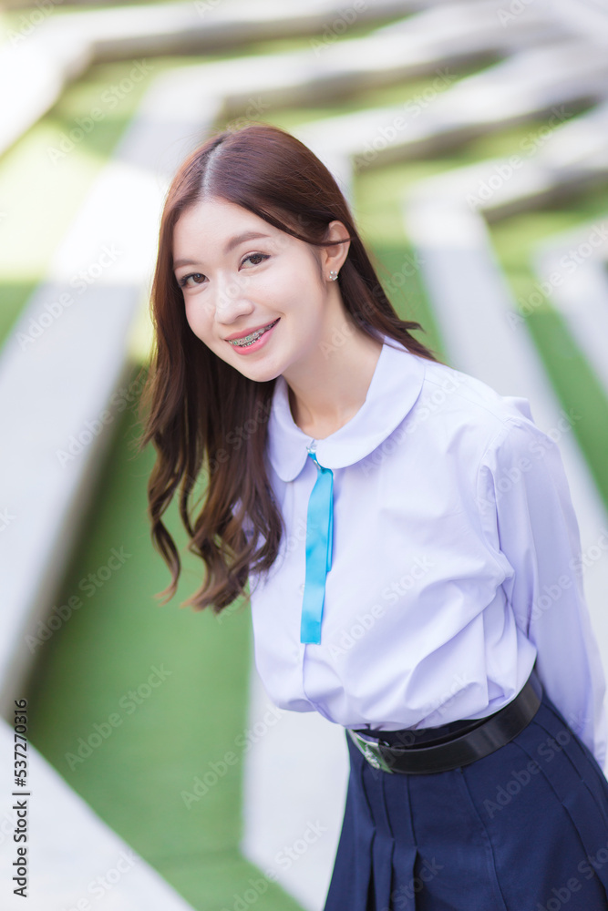 Beautiful Asian high school student young girl in the school uniform with braces on her teeth stands and smiles confidently while she looks at the camera happily with the building in the background. Beautiful Asian high school student young girl in the school uniform with braces on her teeth stands and smiles confidently while she looks at the camera happily with the building in the background.