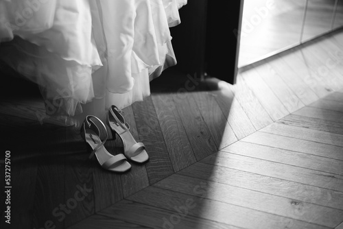 scarpa di matrimonio, vicino al vestito da sposa, dentro ad una camera con pavimento in legno. scarpe alla moda e chic