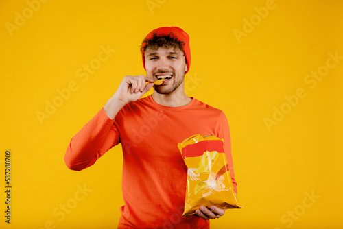 Young happy man holds puts chip in his mouth and holds pack of chips in another, isolated yellow background