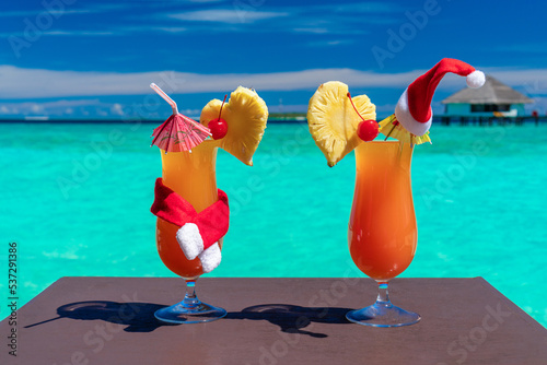 Two bright orange cocktails decarated with santa claus hat and scarf on exotic beach and blue ocean background, nobody.  photo