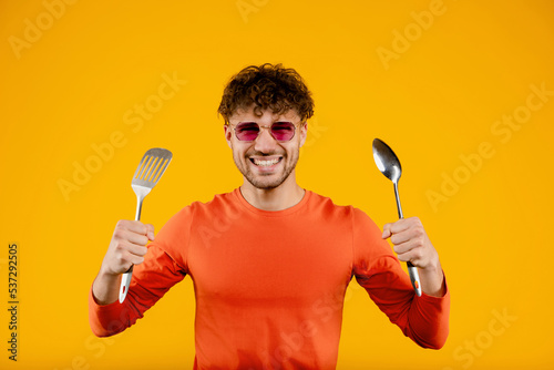 Young man in glasses holds kitchen spatula in one hand and spoon in another , isolated yellow background