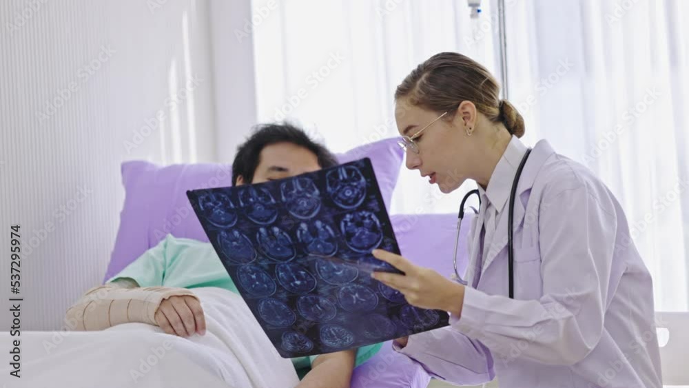 Caucasian female doctor explaining brain X-ray film to Asian male patient listening in hospital bed. Care by expert doctors. Diagnosis of neurodegenerative diseases a blood vessel in brain or cancer Caucasian female doctor explaining brain X-ray film to Asian male patient listening in hospital bed. Care by expert doctors. Diagnosis of neurodegenerative diseases a blood vessel in brain or cancer