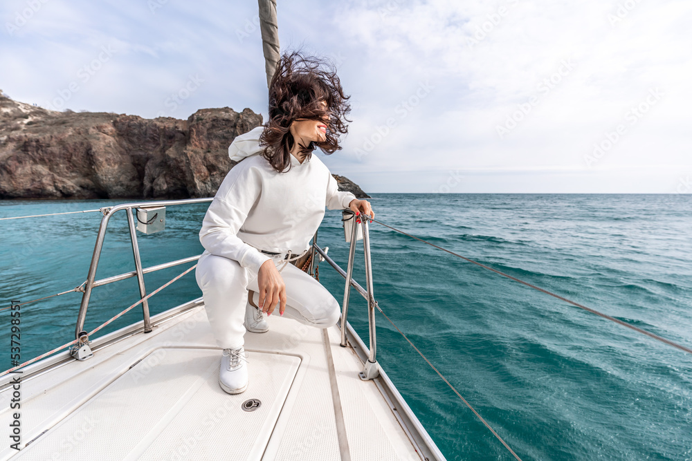 Fototapeta premium A woman sits on the bow of a yacht on a sunny summer day, the breeze develops her hair, a beautiful sea is in the background