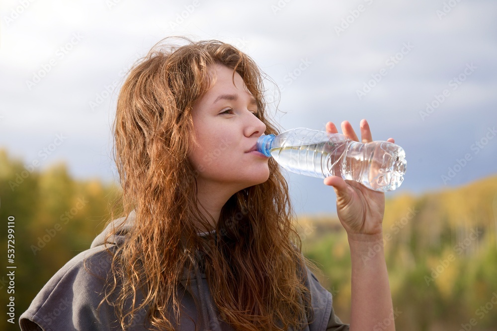 portrait photo of young thirsty woman, beautiful girl is drinking pure fresh water from plastic bottle outdoors on blue sky and autumn forest or park background at sunny day - obrazy, fototapety, plakaty 