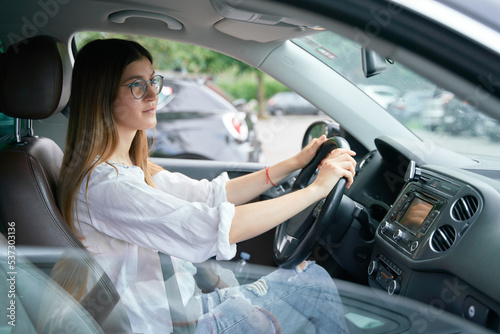 Side view of the beautiful young happy smiling woman driving her car and looking at the road. Portrait of the positive woman wearing glasses keeping steering wheel in the car