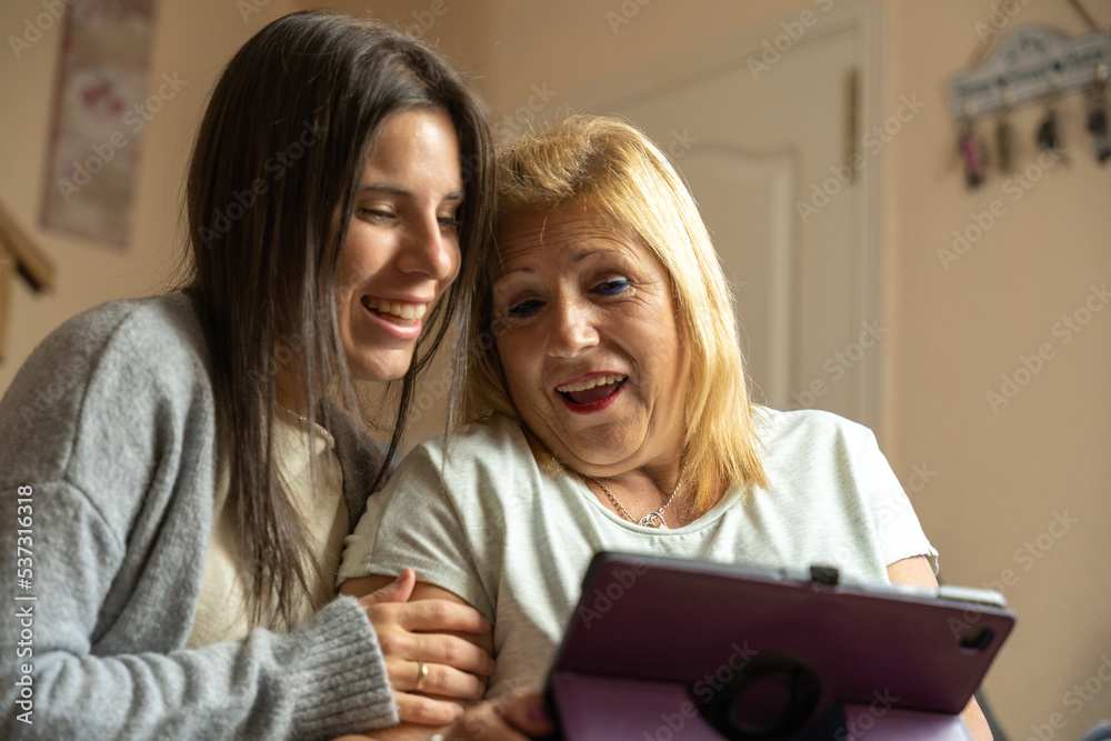 Daughter showing her mother something on the tablet and the mother is surprised while they are happily sitting on the sofa at home. Daughter showing her mother something on the tablet and the mother is surprised while they are happily sitting on the sofa at home.