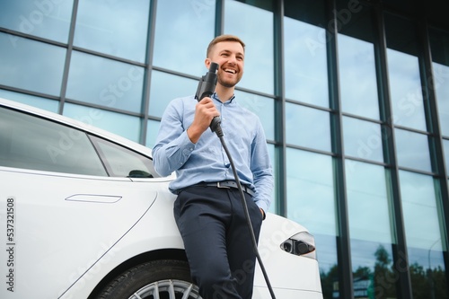 portrait of young handsome bearded man in casual wear, standing at the charging station and holding a plug of the charger for an electric car. Eco electric car concept.