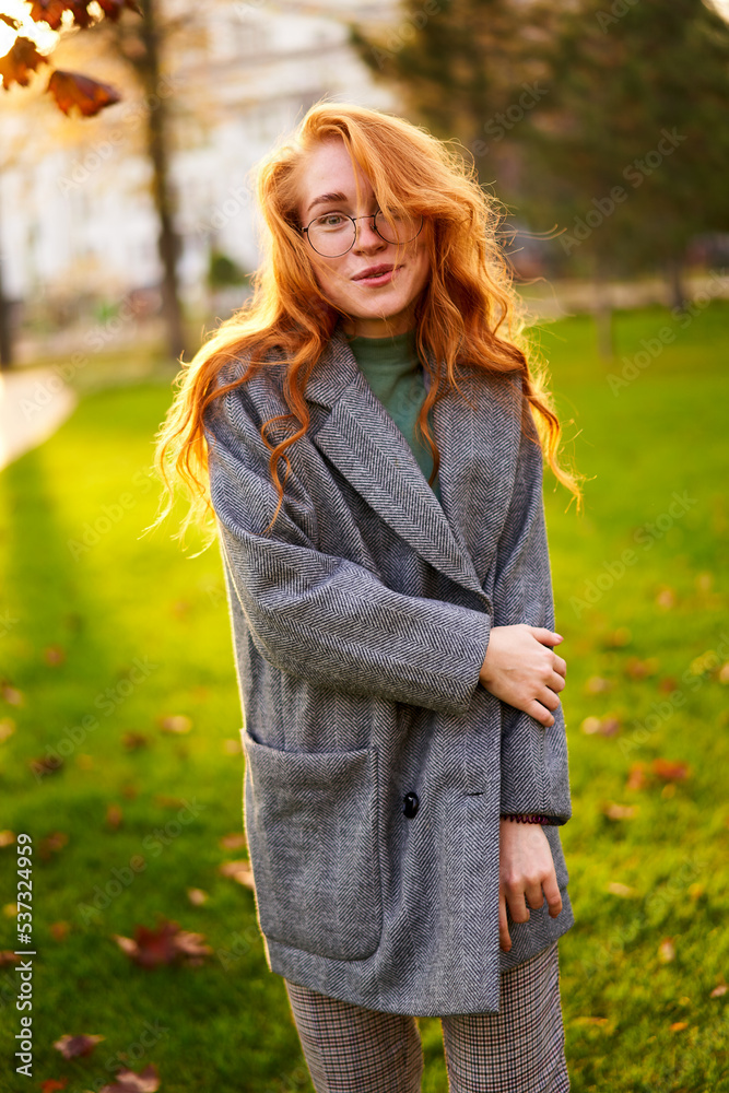 Redhead smiling pretty woman walking in park on sunny autumn day. Elegant female poses in stylish authentic outfit has fun laughing on green lawn with vivid foliage, leaves. Girl enjoys fall season.