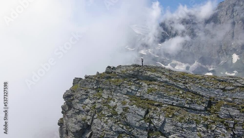 aerial drone movement around the peak of rocky mountains with human hiker standing at the top of the mountain surrounded by clouds