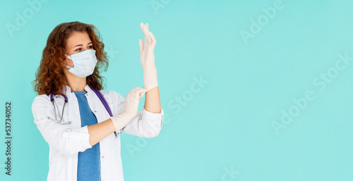 Professional young female physician, doctor in medical mask and white coat, put on rubber gloves for examination, isolated on blue background. Covid-19, coronavirus disease, healthcare workers concept