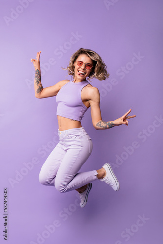Full length funny young caucasian lady winks with tongue sticking out jumping on purple background. Blonde woman wavy hair wears casual clothes. Happiness summer lifestyle concept.