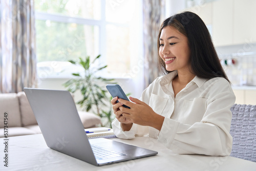 Young Asian woman using mobile phone while working at home with laptop