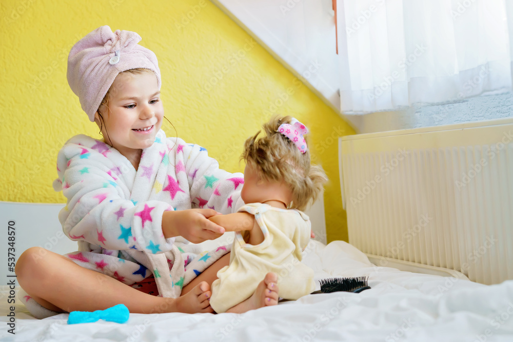 Cute smiling little girl child playing with her doll at home. Preschool child in bathrobe after taking bath brushing hairs of toy doll. Happy kid. Cute smiling little girl child playing with her doll at home. Preschool child in bathrobe after taking bath brushing hairs of toy doll. Happy kid.