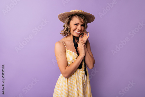 Beautiful young caucasian woman smiles with her teeth looking at camera on purple background. Blonde wavy hair wears summer dress and hat. Emotion concept