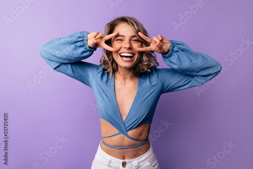Positive young caucasian woman laughing looking at camera showing peace gesture near face standing in studio. Blonde with wavy hair wears blue blouse. Lifestyle, different emotions, leisure concept.