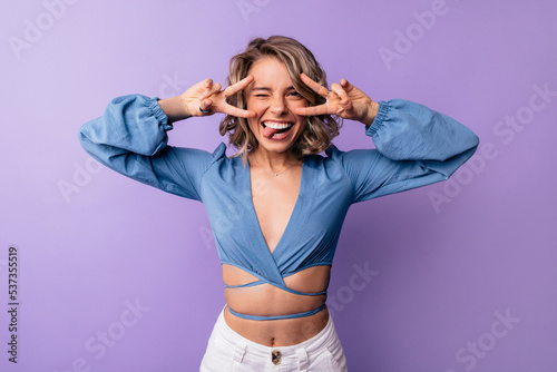 Funny young caucasian girl winks, shows tongue and peace gesture on purple background. Blonde looks at camera, wears blue blouse. Mood concept