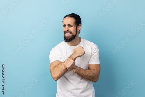 Portrait of sick man with beard wearing white T-shirt standing with grimace of pain, massaging sore wrist, suffering hand injury or sprain. Indoor studio shot isolated on blue background.