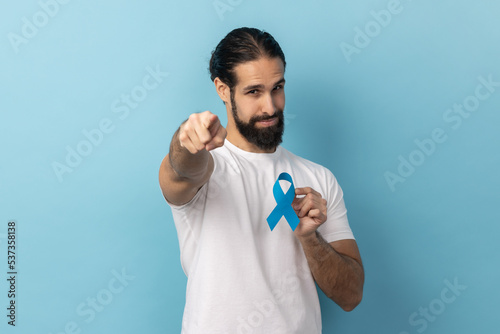 Portrait of responsible handsome man with beard wearing white T-shirt holding blue awareness, disease symbol, pointing to camera, support. Indoor studio shot isolated on blue background. photo