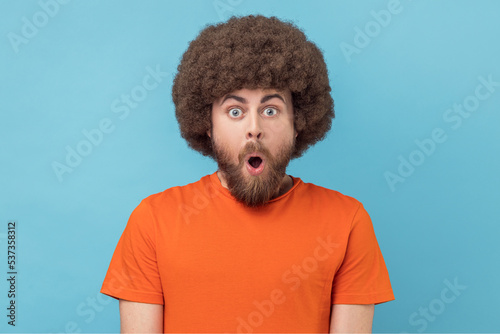Portrait of amazed man with Afro hairstyle wearing orange T-shirt standing with mouth open in surprise, has shocked expression, hears unbelievable news. Indoor studio shot isolated on blue background.