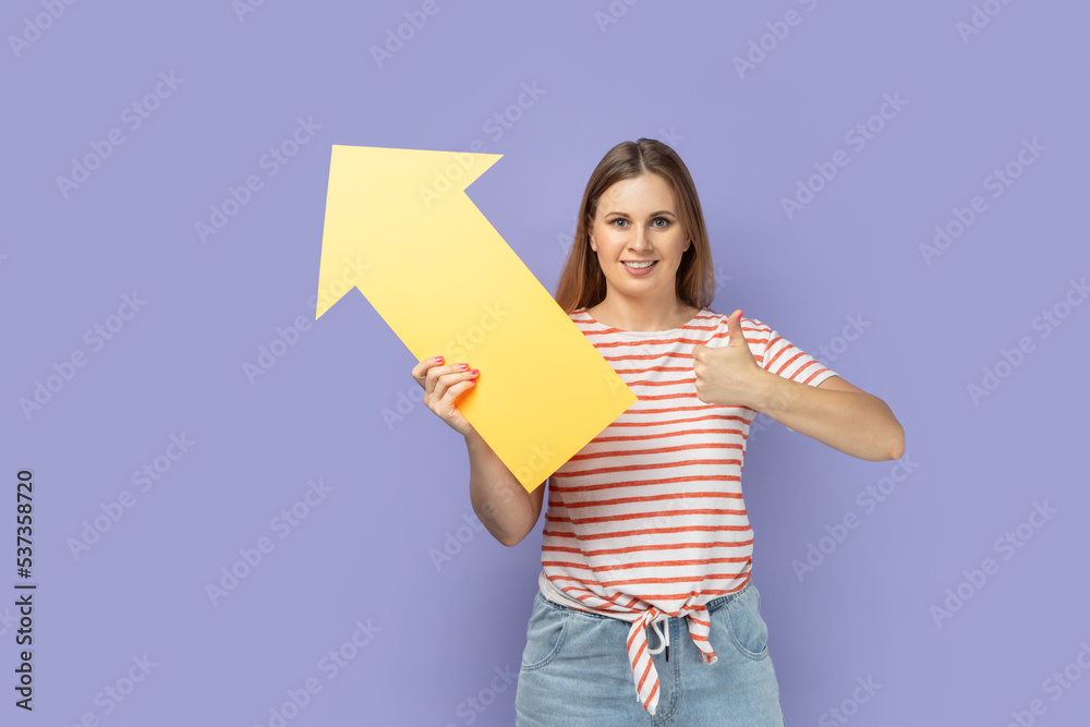 Portrait of pretty smiling blond woman wearing striped T-shirt holding big yellow arrow pointing aside and showing thumb up, like gesture. Indoor studio shot isolated on purple background. Portrait of pretty smiling blond woman wearing striped T-shirt holding big yellow arrow pointing aside and showing thumb up, like gesture. Indoor studio shot isolated on purple background.
