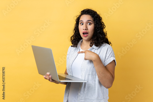 Portrait of amazed astonished woman with dark wavy hair standing with laptop in hands and pointing at display, looking with big eyes and open mouth. Indoor studio shot isolated on yellow background. © khosrork