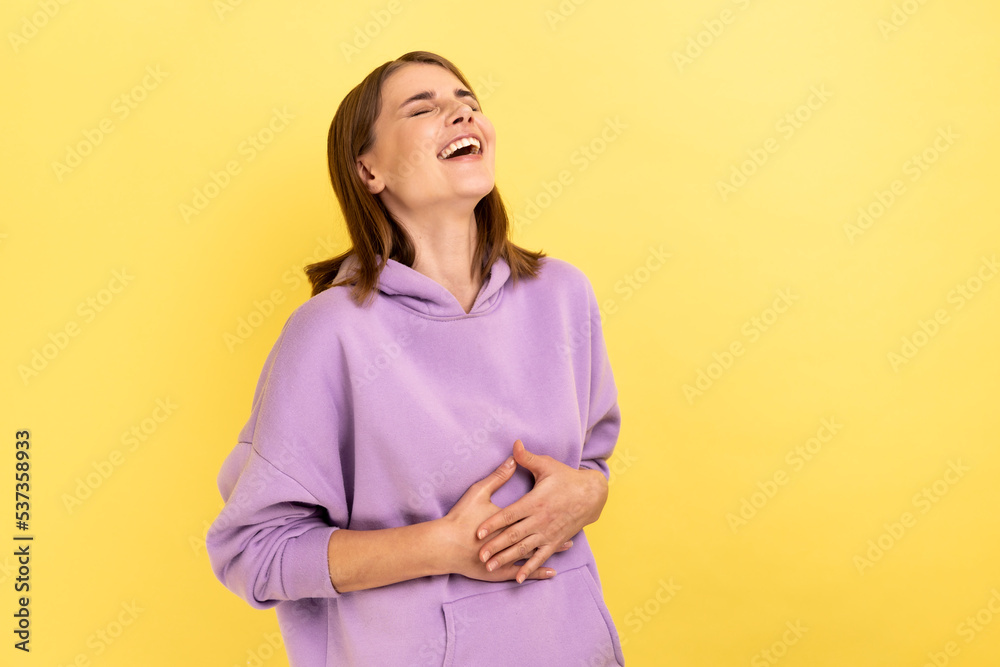 Obraz premium Portrait of happy woman with dark hair holding belly, laughing, hearing funny joke or anecdote, being in good mood, wearing purple hoodie. Indoor studio shot isolated on yellow background.