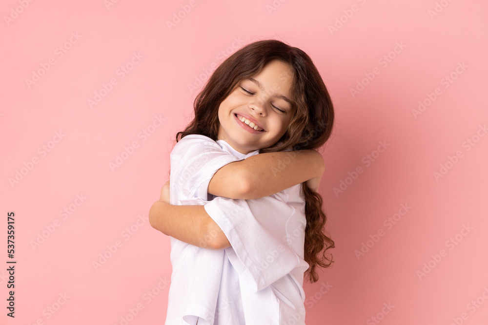 Portrait of satisfied happy little girl wearing white T-shirt embracing herself, standing with eyes closed from pleasure and happiness. Indoor studio shot isolated on pink background. Portrait of satisfied happy little girl wearing white T-shirt embracing herself, standing with eyes closed from pleasure and happiness. Indoor studio shot isolated on pink background.