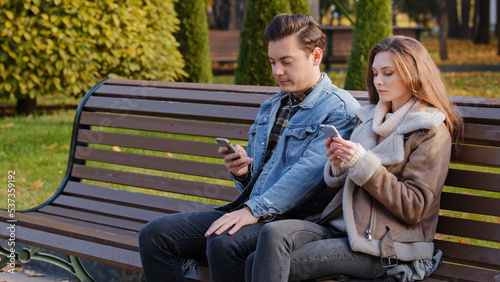 Caucasian couple two young people partners sit on bench use phone cell gadgets scroll internet web pages check social networks show images each other spend time in city park suffer of mobile addiction