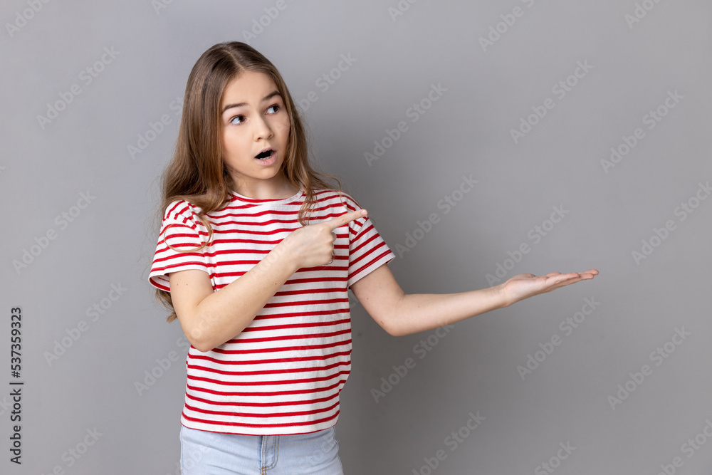 Portrait of surprised little girl wearing striped T-shirt pointing at copy space on her palm, empty place for idea presentation, product advertising. Indoor studio shot isolated on gray background. Portrait of surprised little girl wearing striped T-shirt pointing at copy space on her palm, empty place for idea presentation, product advertising. Indoor studio shot isolated on gray background.