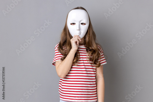 Photo Portrait of anonymous unknown little girl wearing striped T-shirt covering her face with white mask, hiding her real personality, anonymity