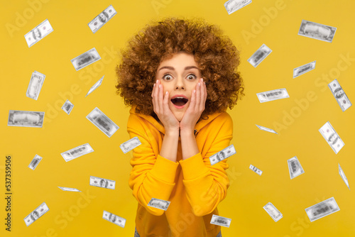 Oh my god, wow. Portrait of woman with Afro hairstyle looking at camera with big eyes, absolutely shocked of money rain falling from up. Indoor studio shot isolated on yellow background.