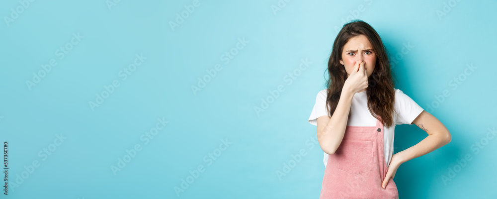 Portrait of bothered cute girl shut nose and frown from bad smell, smelling terrible reek, something stink, standing against blue background - obrazy, fototapety, plakaty 