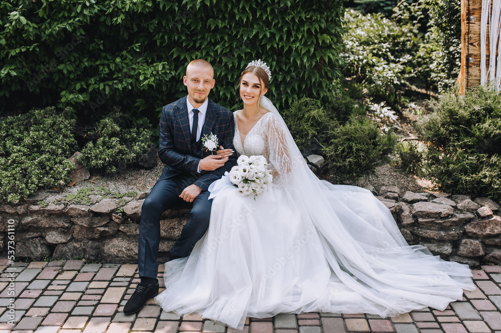 A stylish, young groom and a beautiful bride in a white dress with a bouquet in their hands, a diadem on their heads, sit gently hugging in a park in nature. Wedding photography of the newlyweds.