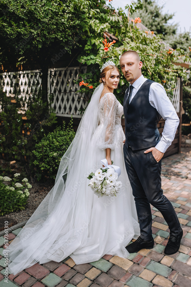 A stylish bearded groom and a beautiful young bride in a white long dress with a diadem on her head with a bouquet are standing in a park, a garden in nature. Wedding photography, portrait.