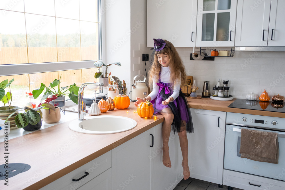 Child decorates the kitchen in home for Halloween. Girl in a witch costume plays with the decor for the holiday - bats, jack lantern, pumpkins. Autumn comfort in house, Scandi-style kitchen, loft