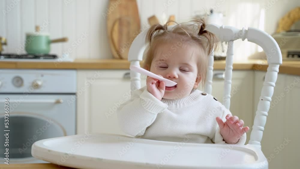 Child baby girl licking plastic spoon while waiting for mother cooking lunch food. Kid makes funny faces. Baby feeding concept Child baby girl licking plastic spoon while waiting for mother cooking lunch food. Kid makes funny faces. Baby feeding concept