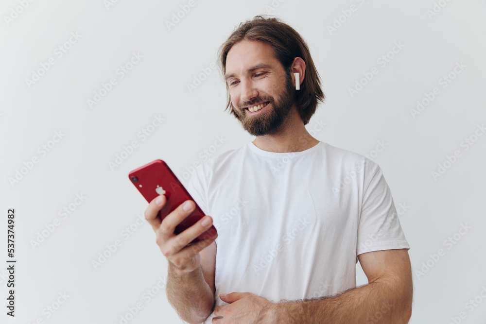 A man with a beard in white T-shirt and jeans looks at his phone flipping through an online social media feed with headphones in his ears listening to a voice message on a white background and smiling A man with a beard in white T-shirt and jeans looks at his phone flipping through an online social media feed with headphones in his ears listening to a voice message on a white background and smiling