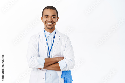 Handsome arab doctor in white coat  cross arms on chest  looks professional  stands over white background