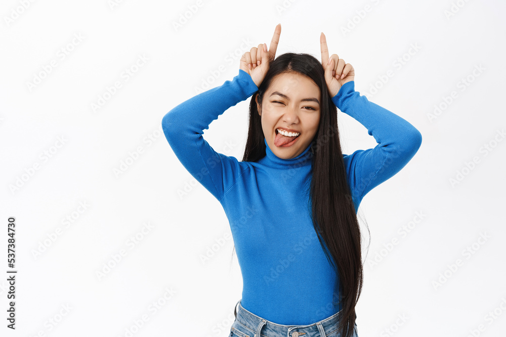 Enthusiastic asian woman shows devil horns, fingers on head, smiles sassy and extends tongue, playful and flirty emotion, white background