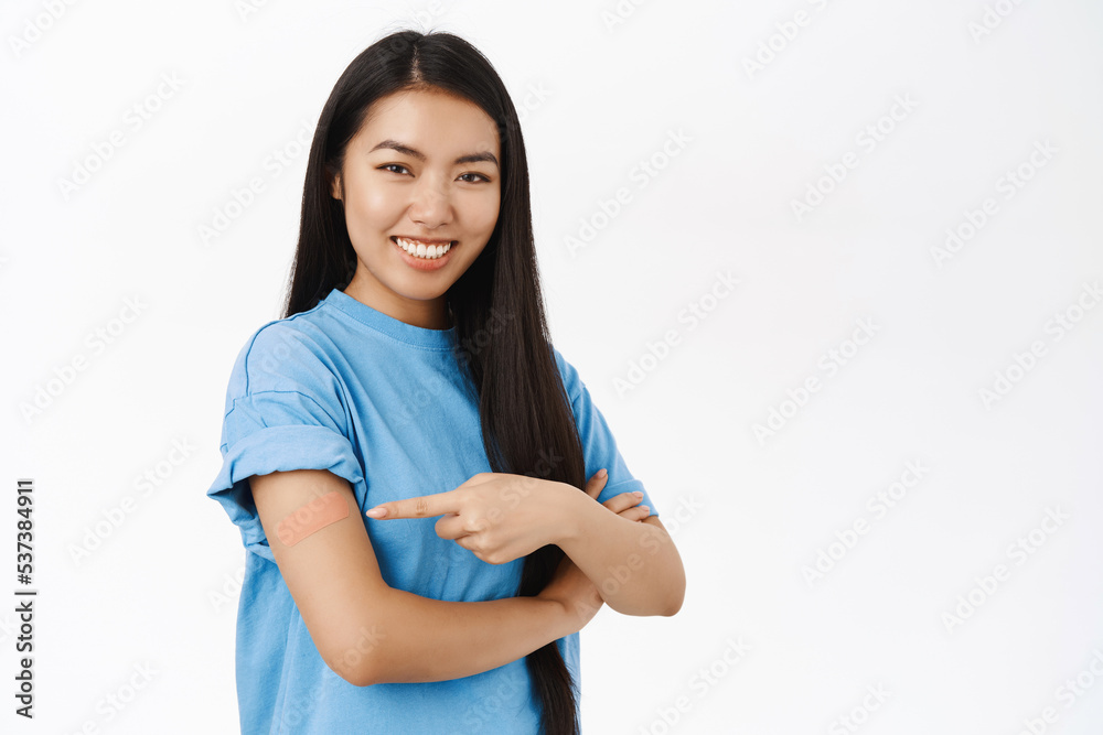 Smiling chinese girl points at shoulder with band aid, promotes vaccination from covid-19, coronavirus vaccine advertisement, white background Smiling chinese girl points at shoulder with band aid, promotes vaccination from covid-19, coronavirus vaccine advertisement, white background
