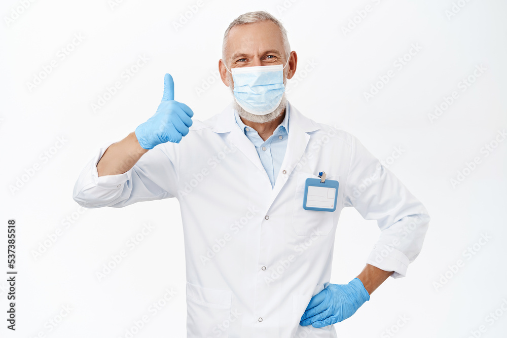 Doctor in medical face mask from covid-19, wearing rubber gloves and white coat, showing thumbs up, recommending hospital or clinic, standing over white background Doctor in medical face mask from covid-19, wearing rubber gloves and white coat, showing thumbs up, recommending hospital or clinic, standing over white background