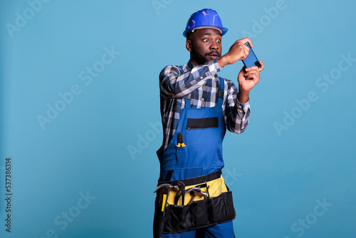 African american builder taking a break at work playing video games on a cell phone. Construction worker wearing work uniform relaxing after a busy day against blue background in studio shot. © DC Studio