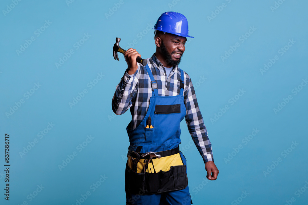 Aggressive builder holding hammer hard wearing blue protective helmet and uniform. African American man at work looking furious stressed after many hours of heavy work without rest. Aggressive builder holding hammer hard wearing blue protective helmet and uniform. African American man at work looking furious stressed after many hours of heavy work without rest.