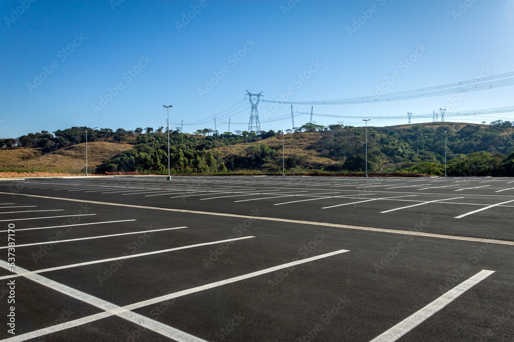 Huge parking lot completely empty. In the background, hill covered with trees and power transmission lines.