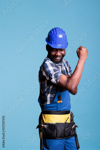 Strong man flexing arm muscles, preparing to work on construction reform with tools. Confident and energetic african american builder pointing biceps and triceps, studio shot against blue background. © DC Studio
