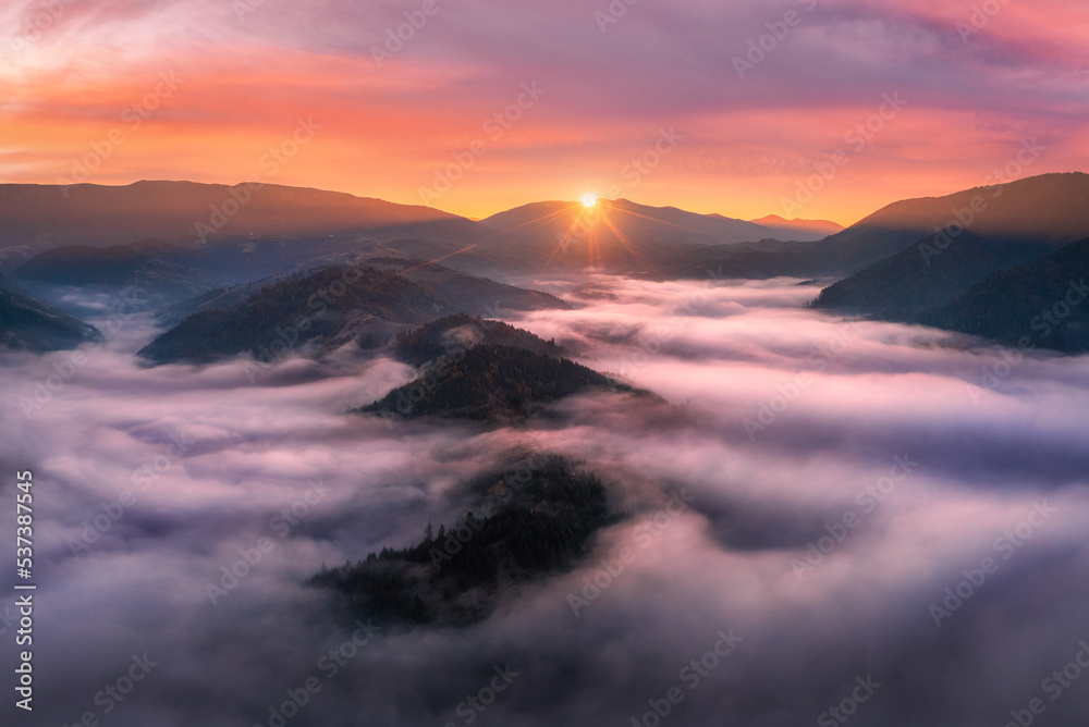 Aerial view of mountains in low clouds at sunrise in autumn. Top view of mountain hills in fog in fall at dawn. Beautiful landscape with ridges, forest, sun, colorful orange sky with pink clouds