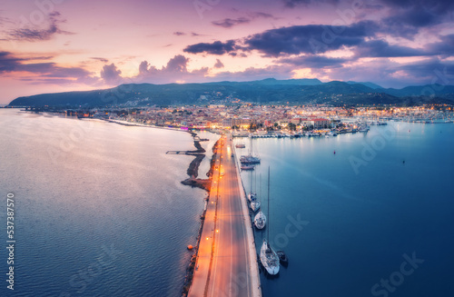 Aerial view of road near sea canal at night in summer in Lefkada island, Greece. Top view of road, blurred cars, boats and yachts, city lights, architecture, mountain and sky with clouds at sunset photo