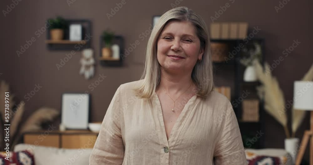 Headshot portrait positive mature woman with healthy smile standing on background of cozy living room, happy middle-aged lady looking to camera, posing for photo at home, enjoying leisure time. Headshot portrait positive mature woman with healthy smile standing on background of cozy living room, happy middle-aged lady looking to camera, posing for photo at home, enjoying leisure time.