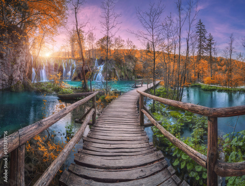 Waterfall and wooden path in orange forest in Plitvice Lakes, Croatia at sunset in autumn. Colorful landscape with trail in park, trees, water lilies, river, pink sky in fall. Trail in woods. Nature