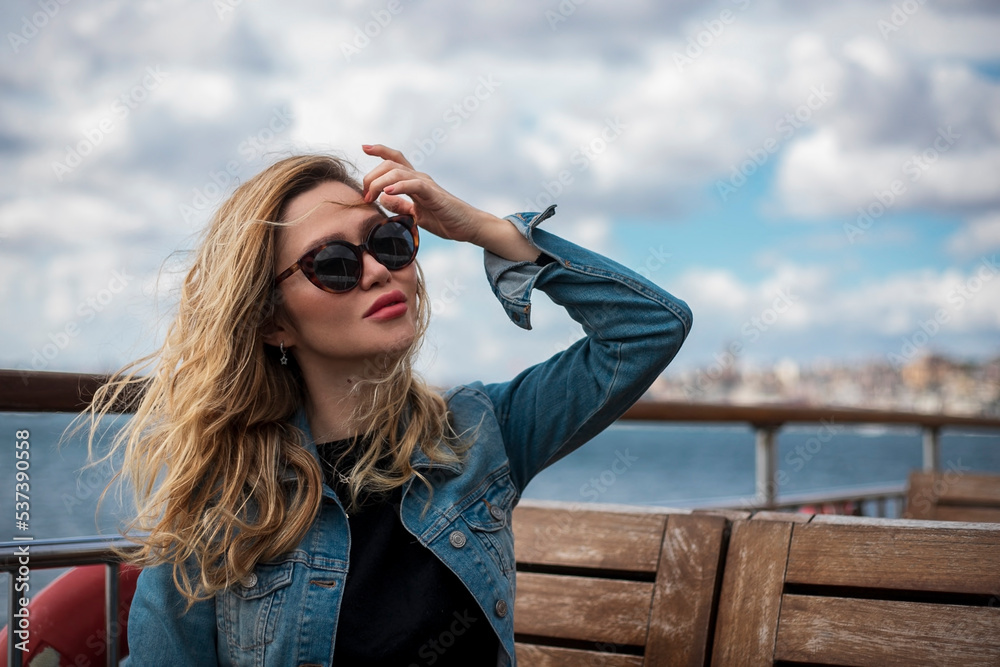 Blonde woman wearing sunglasses, sitting on the deck of the ferry, holding her blowing hair in the wind. Bosphorus, Turkey. Blonde woman wearing sunglasses, sitting on the deck of the ferry, holding her blowing hair in the wind. Bosphorus, Turkey.